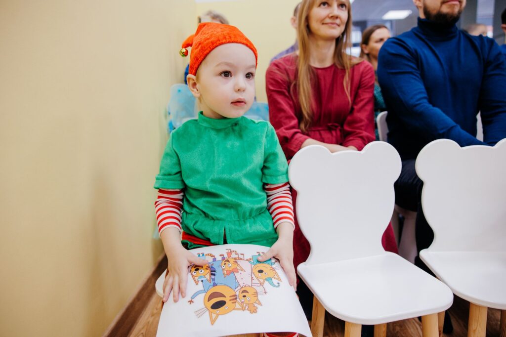 A young child dressed as an elf sits at a Christmas event indoors.